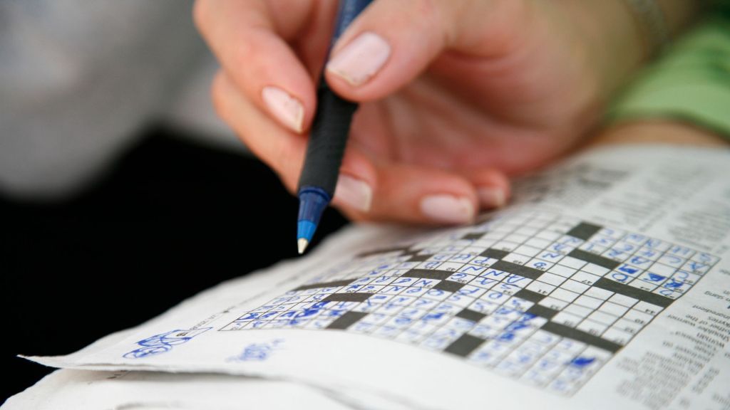 Image of a hand holding a blue pen above a crossword puzzle in a newspaper